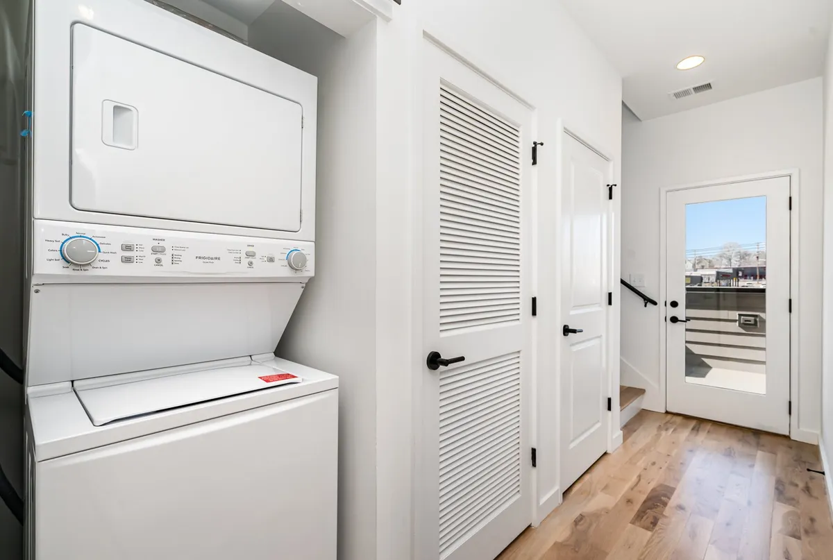 This image features a stacked washer and dryer unit in a practical nook with white double doors, wood flooring, and a window allowing in natural light. The space is functional and clean, evoking a sense of organization and convenience for guests handling daily chores during their stay.
