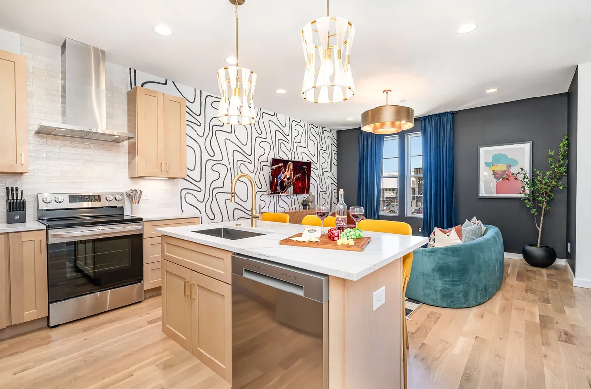 A vibrant kitchen and living area where a kitchen island is adorned with wine and snacks, bold patterned walls create visual interest, and large windows drench the area in natural sunlight. Colorful furniture and blue curtains add a playful, welcoming touch, ideal for entertaining or relaxing.
