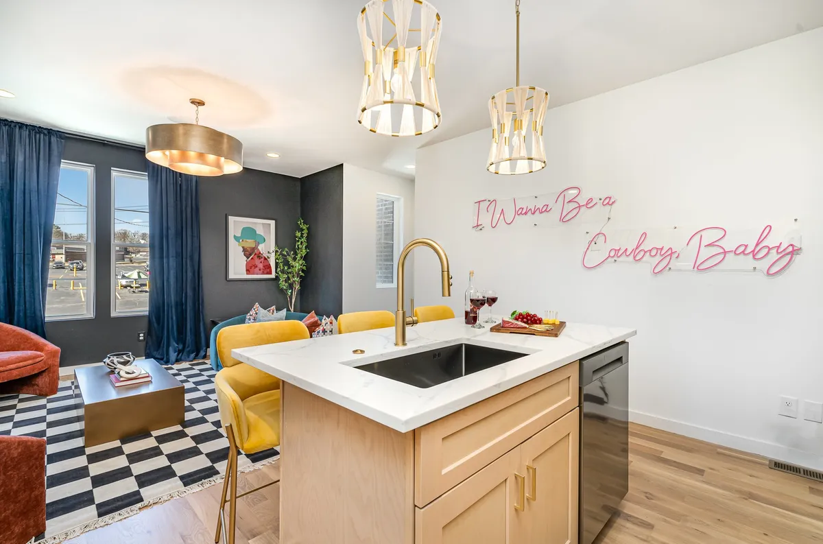 A vibrant kitchen setting is shown, featuring a kitchen island, colorful chairs, a neon sign, and large windows. The area is open and bright, inviting for both meal preparation and casual gatherings. The modern design signals attention to detail and enhances the welcoming atmosphere for groups and families.