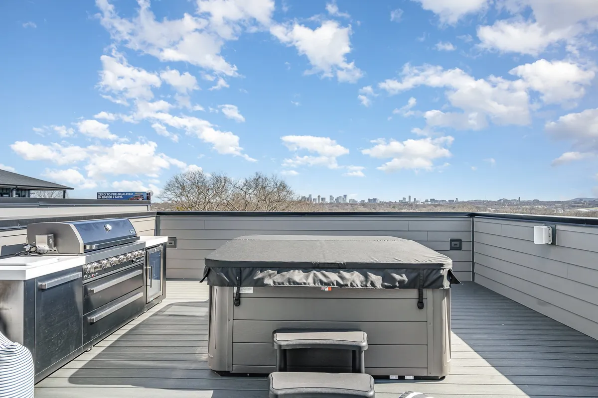 A rooftop patio set against the city skyline, featuring a bubbling hot tub, an outdoor grill, and comfortable seating for gatherings. The city lights create a vibrant backdrop, suggesting possibilities for both lively social events and tranquil evenings above the city.