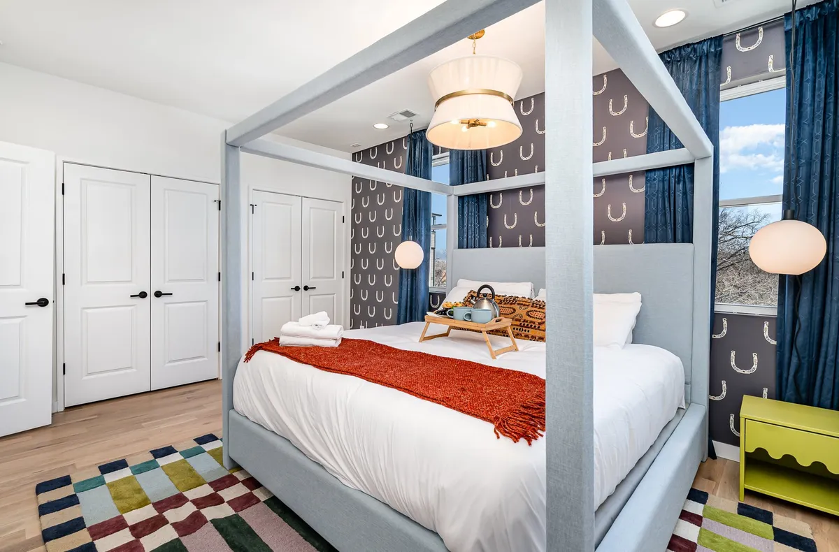 A stylish bedroom featuring a canopy king bed, a vibrant and colorful rug, decorative pillows, and modern lighting fixtures. The room exudes a cozy yet upscale vibe, with thoughtful design elements that promote relaxation and comfort.